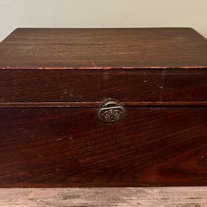 May include: A rectangular wooden box with a dark brown finish. The box has a metal clasp and is likely used for storage. The wood grain is visible, and the box appears to be vintage or antique.