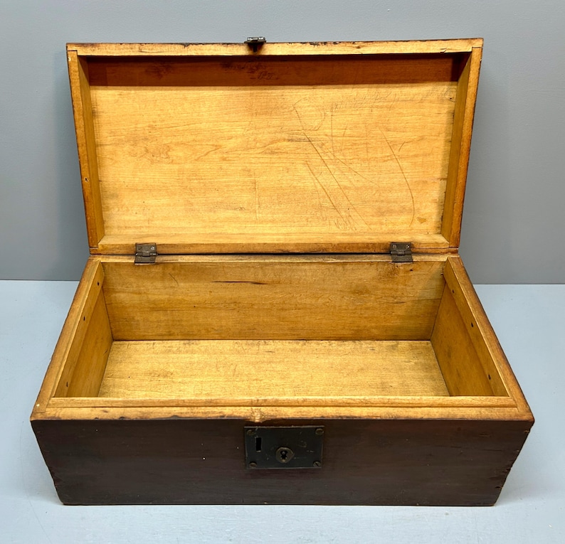 May include: An open, antique wooden chest with a dark brown exterior and a light brown interior. The lid is open, revealing the empty space inside. The chest has a metal latch and hinges. The wood grain is visible.