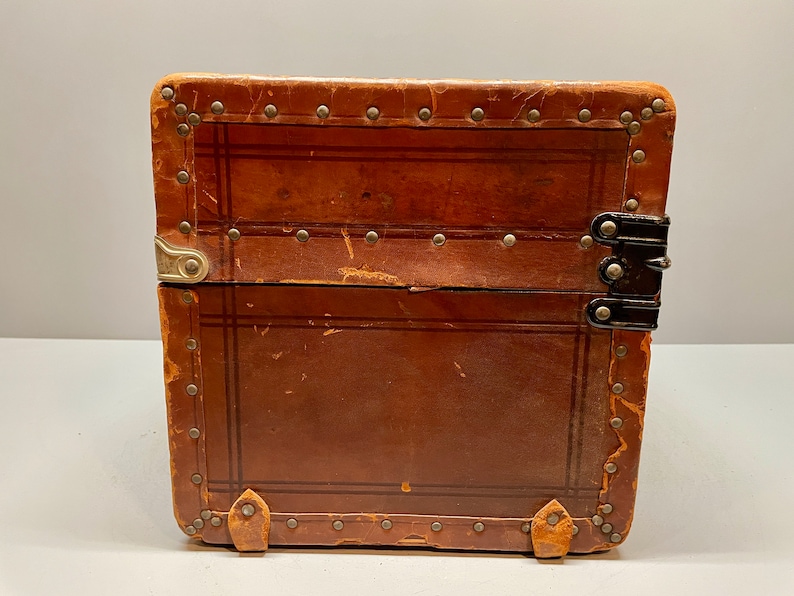May include: A vintage brown leather suitcase with metal accents and a black latch. The suitcase has a worn and distressed look, suggesting it has been well-loved and traveled with.