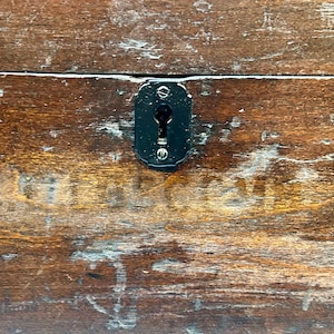May include: Close-up of a weathered wooden surface with a dark, oval-shaped keyhole plate. The wood has a rich, brown color with visible grain and signs of wear, suggesting age and use. The keyhole plate is black and secured with small screws.