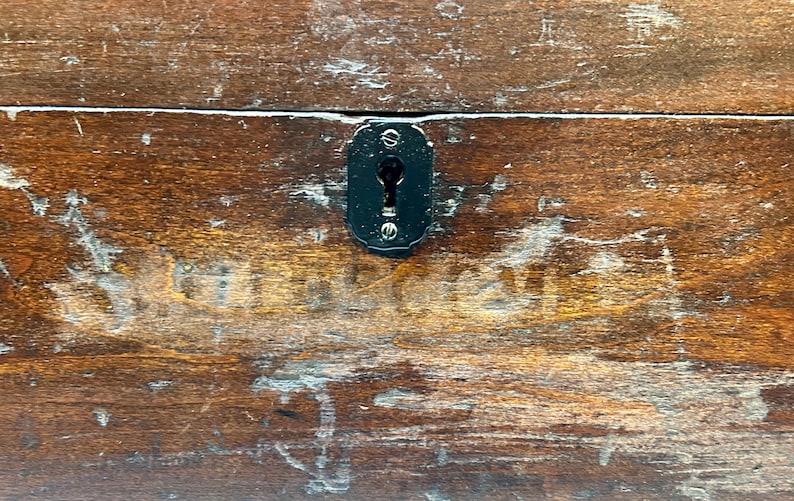 May include: Close-up of a weathered wooden surface with a dark, oval-shaped keyhole plate. The wood has a rich, brown color with visible grain and signs of wear, suggesting age and use. The keyhole plate is black and secured with small screws.