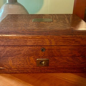 May include: A rectangular wooden box with a dark brown wood grain finish. The box features a hinged lid with a small rectangular brass plate on top, a central keyhole on the front panel, and a brass pull handle centered on the lower front section.