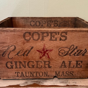 May include: Vintage wooden crate with the text "COPE'S Red Star GINGER ALE Taunton, MASS." in black lettering and a red star. The crate is rectangular, with a weathered, aged appearance, and is likely from the early 20th century.