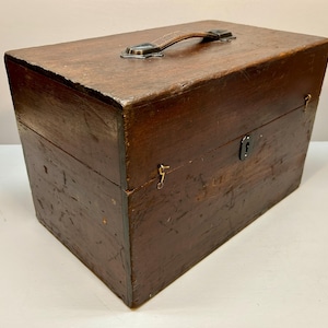 May include: A vintage wooden box with a leather handle and metal hardware. The box is rectangular, with a dark brown finish and visible wood grain. It features a small lock and hinges, suggesting it may have been used for storage or transport.