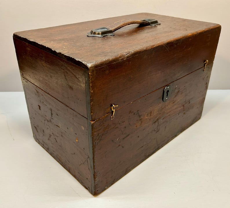 May include: A vintage wooden box with a leather handle and metal hardware. The box is rectangular, with a dark brown finish and visible wood grain. It features a small lock and hinges, suggesting it may have been used for storage or transport.
