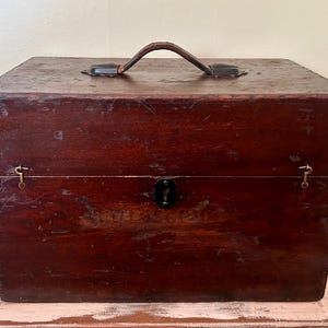 May include: A vintage wooden box with a dark brown finish. The box has a leather handle on top and metal hardware, including a lock and hinges. The box is rectangular and appears to be well-worn, suggesting age and use. It sits on a light-colored surface.