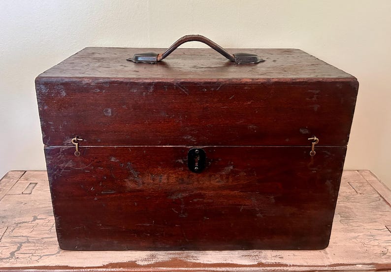 May include: A vintage wooden box with a dark brown finish. The box has a leather handle on top and metal hardware, including a lock and hinges. The box is rectangular and appears to be well-worn, suggesting age and use. It sits on a light-colored surface.