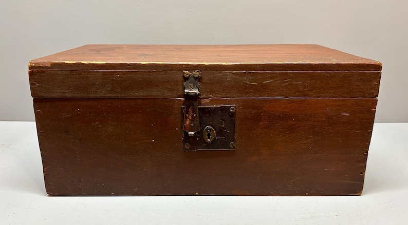 May include: A rectangular wooden chest with a hinged lid and a metal clasp. The chest is stained a dark brown color, with visible wood grain. The metal clasp and keyhole plate show signs of age and wear.