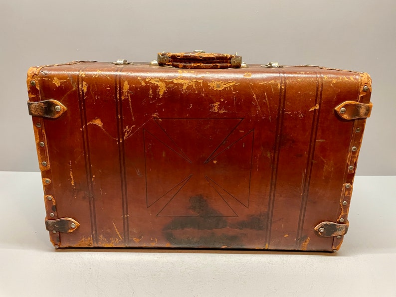 May include: A vintage brown leather suitcase with a decorative cross pattern and metal accents. The suitcase has a worn and weathered look, suggesting it has been well-traveled.
