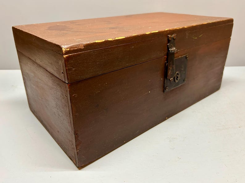 May include: A rectangular wooden box with a hinged lid, painted in a dark brown color. The box features a metal latch and keyhole on the front. The wood shows signs of wear, with some paint chipping.