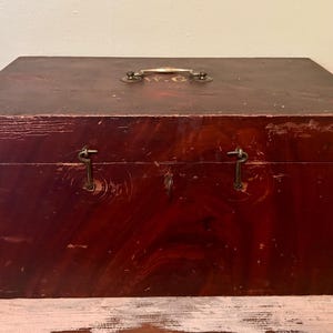 May include: A rectangular, antique wooden box with a dark reddish-brown finish. The box has a metal handle and two metal clasps. The wood shows signs of wear, adding to its vintage appeal. The box is likely used for storage.