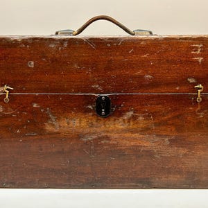 May include: A vintage wooden toolbox with a leather handle and brass hardware. The box is rectangular with a hinged lid and a lock. The wood has a rich, dark brown color with visible wear and tear, suggesting age and use.