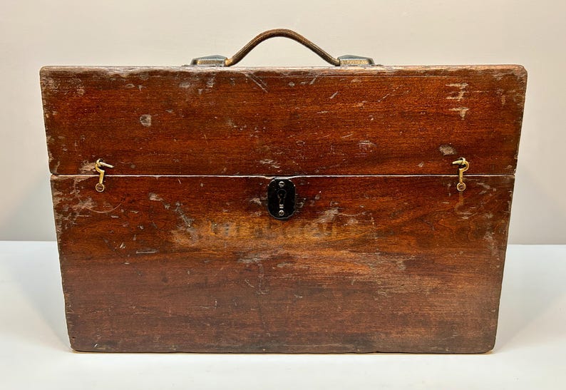 May include: A vintage wooden toolbox with a leather handle and brass hardware. The box is rectangular with a hinged lid and a lock. The wood has a rich, dark brown color with visible wear and tear, suggesting age and use.