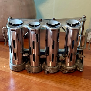 May include: A vintage metal coin dispenser featuring four vertical cylindrical tubes for sorting and holding coins. The silver-toned device has open tops, vertical slots along each tube, and small levers at the base of each section for dispensing.