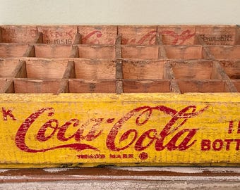 May include: A rectangular wooden Coca-Cola crate with a yellow exterior and red lettering that reads Drink Coca-Cola In Bottles. The crate features internal dividers for twenty-four bottles and has metal corner reinforcements on each side.