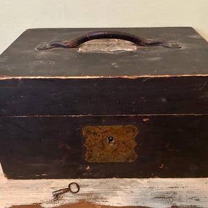 May include: A vintage black wooden box with a leather handle and metal accents. The box has a keyhole and a small key is lying in front of it. The box appears to be aged and worn, suggesting it is an antique.