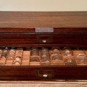 May include: A dark brown wooden storage cabinet with two drawers, featuring a metal label plate that reads Universal Watch Crystals. The bottom drawer is open, revealing multiple internal compartments filled with various clear glass watch crystals.