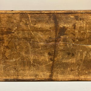 May include: A rectangular wooden box with a distressed finish. The box is made of light brown wood with visible grain and numerous scratches, giving it a rustic appearance. The box has a simple, classic design.