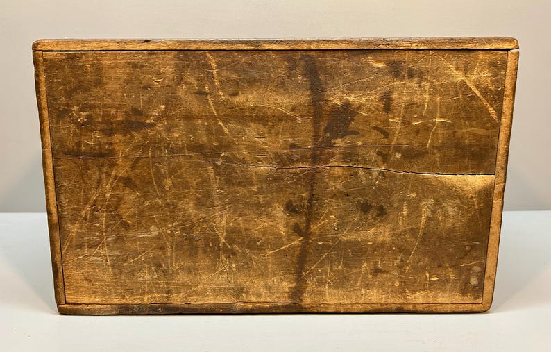 May include: A rectangular wooden box with a distressed finish. The box is made of light brown wood with visible grain and numerous scratches, giving it a rustic appearance. The box has a simple, classic design.
