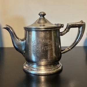 May include: A small, silver-colored teapot with a lid and handle. The words "ST. FRANCIS HOSPITAL" are engraved on the side. The teapot has a spout and sits on a small base. The teapot is likely vintage.