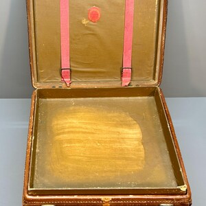 May include: Open antique brown leather suitcase with a brass clasp and hardware. The interior features a light brown lining, pink straps, and a circular emblem. The suitcase is rectangular and appears to be vintage.