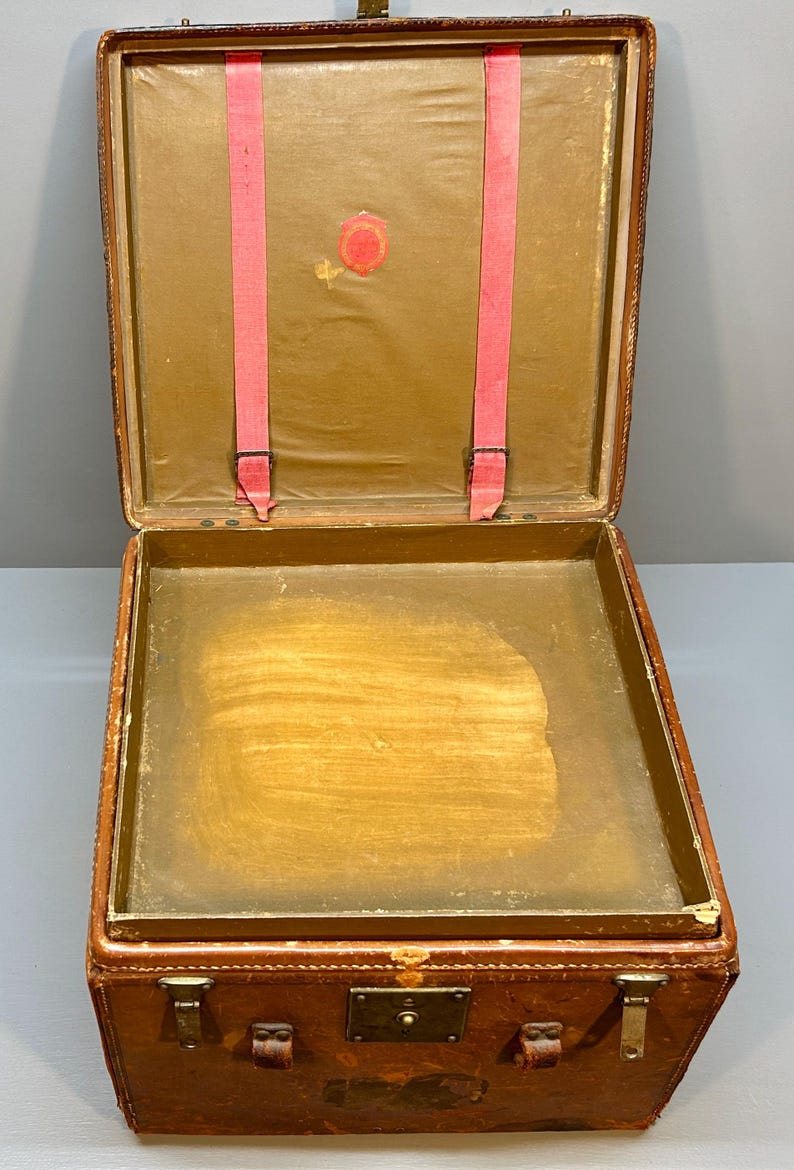 May include: Open antique brown leather suitcase with a brass clasp and hardware. The interior features a light brown lining, pink straps, and a circular emblem. The suitcase is rectangular and appears to be vintage.