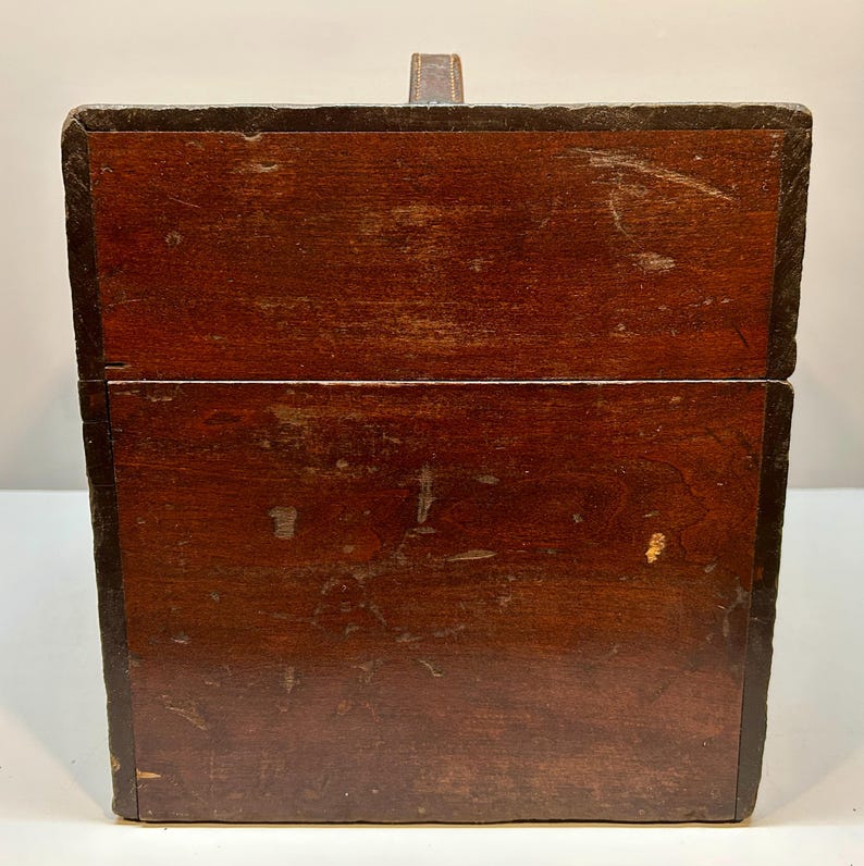 May include: A vintage wooden box with a dark brown finish and a leather handle. The box has a rectangular shape with a hinged lid and appears to be well-worn, showing signs of age and use. The edges are framed with a darker material.