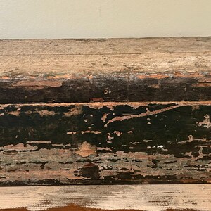 May include: A rectangular wooden box with a hinged lid. The box has a distressed, weathered appearance with peeling paint in shades of black, green, and faded pink. The wood grain is visible, adding to its rustic charm.