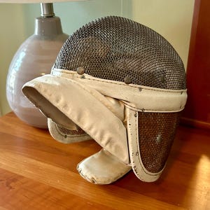 May include: A white and brown fencing mask with a wire mesh face guard. The mask has a white leather strap and a white leather chin guard.
