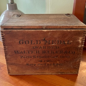 May include: A vintage wooden box with a hinged lid. The box is stained a medium brown and features the text "GOLD MEDAL AWARDED WALTER BAKER & Co. Paris Expositions 1900" in black lettering. The text "Keep cool and dry" is also visible.
