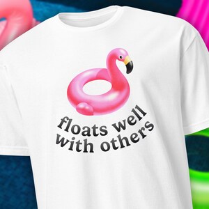 May include: White t-shirt with a pink flamingo inflatable graphic and the text "floats well with others". The flamingo has a black beak and yellow accents. A summer-themed apparel item.
