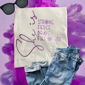 May include: A cream-colored t-shirt with the words "Strong, Fierce, Brave, Full of Fire" in purple glitter lettering. The shirt is paired with light blue jeans and black sunglasses. The background has a purple smoky effect.