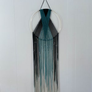 May include: A macrame wall hanging with a gold hoop and black and teal yarn. The yarn is ombre, fading from black to teal to white.