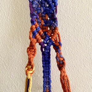 Pode incluir: Um close-up de um suporte de planta de macramé com cordões azuis, roxos, laranja e amarelos. Os cordões são amarrados juntos em um padrão complexo.