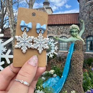 May include: A pair of snowflake earrings with light blue bows. The earrings are white with silver glitter accents. The earrings are displayed on a brown card. The background includes a stone building, a statue of a woman, and a garden.