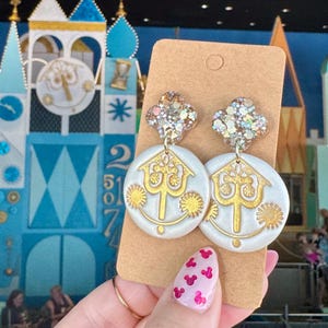 May include: A pair of dangle earrings featuring a clover-shaped top with multi-colored glitter and a round white base with a gold-colored clock face design. The earrings are attached to a rectangular brown cardboard display card.