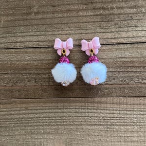 May include: A pair of pink bow earrings with a white fluffy pom-pom design. The earrings have a pink bow at the top, a pink beaded section, and a white, fluffy pom-pom at the bottom. The earrings are displayed on a wooden surface.
