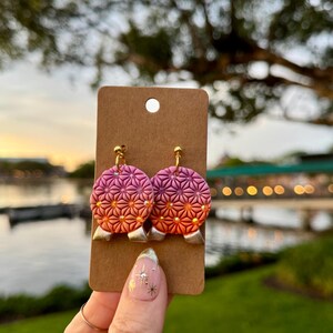 May include: Handmade earrings with a geometric pattern in pink, orange, and gold. The earrings are presented on a brown card. The background is a blurred view of water and greenery. The earrings are a unique accessory.