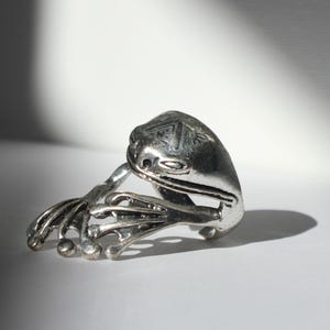 May include: A silver-toned metal ring shaped like a frog. The ring features a detailed frog head with etched markings on the top and extended front legs with splayed toes that wrap around the finger band. The surface has a polished metallic finish.