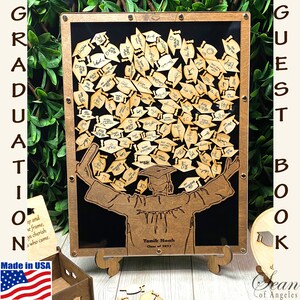 Op de afbeelding: Een houten gastenboek met een afstudeerthema. Het boek toont een silhouet van een afgestudeerde die een diploma vasthoudt met een pet en toga. De achtergrond is zwart en het boek is ingelijst met hout. Het boek is versierd met kleine houten afstudeerpetten die gasten kunnen signeren. De tekst "GUEST BOOK" staat aan de rechterkant van de lijst. De tekst "Yanik Moah Class of 2023" staat onderaan de lijst.