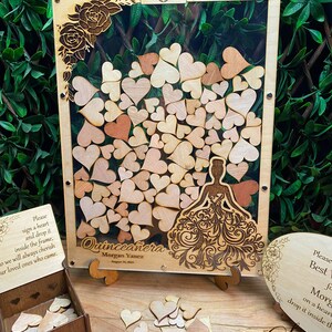 May include: Wooden Quincea&ntilde;era guest book with a clear acrylic front, filled with wooden hearts. The frame features a laser-cut design of a quincea&ntilde;era dress and floral accents. Includes a wooden box and a heart-shaped sign.