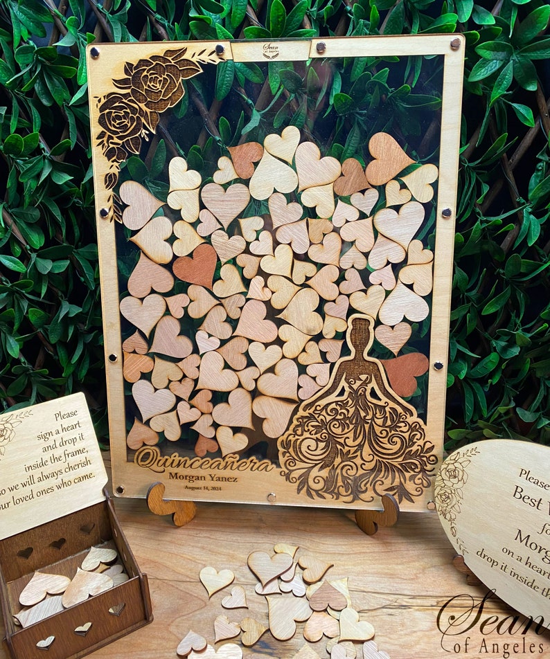 May include: Wooden Quincea&ntilde;era guest book with a clear acrylic front, filled with wooden hearts. The frame features a laser-cut design of a quincea&ntilde;era dress and floral accents. Includes a wooden box and a heart-shaped sign.