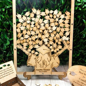 Op de afbeelding: Een houten afstudeerhoedboom met een transparant frame, een klein houten doosje en een bord met de tekst "Schrijf je beste wensen voor Camilla, klas van 2023, op een chip en laat het in het frame vallen".
