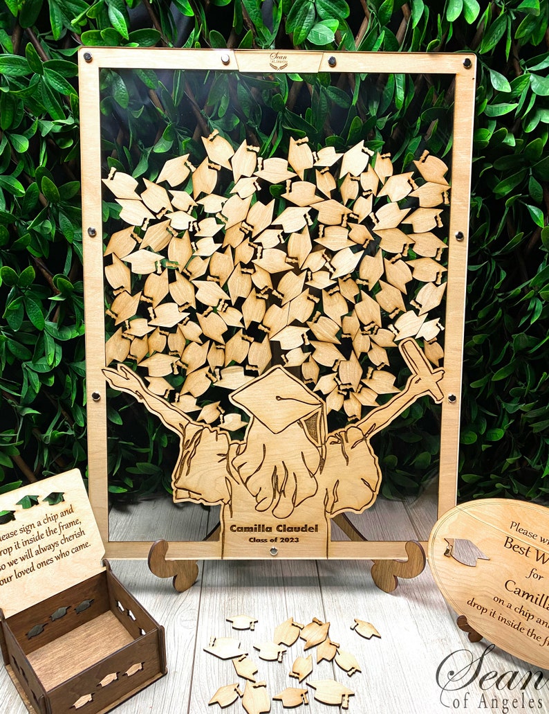 Op de afbeelding: Een houten afstudeerhoedboom met een transparant frame, een klein houten doosje en een bord met de tekst "Schrijf je beste wensen voor Camilla, klas van 2023, op een chip en laat het in het frame vallen".