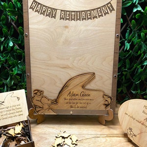 May include: A wooden sign with the text "Happy Retirement" and a fishing theme. The sign has a cutout of a fisherman holding a fishing rod with a fish on the line. The sign also has a cutout of a frog. The sign is on a wooden stand. There is a box of wooden fish shapes and a heart-shaped sign with a fishing theme.