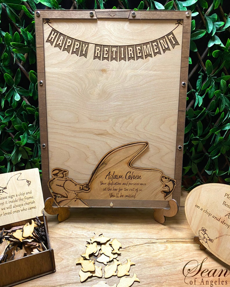May include: A wooden sign with the text "Happy Retirement" and a fishing theme. The sign has a cutout of a fisherman holding a fishing rod with a fish on the line. The sign also has a cutout of a frog. The sign is on a wooden stand. There is a box of wooden fish shapes and a heart-shaped sign with a fishing theme.