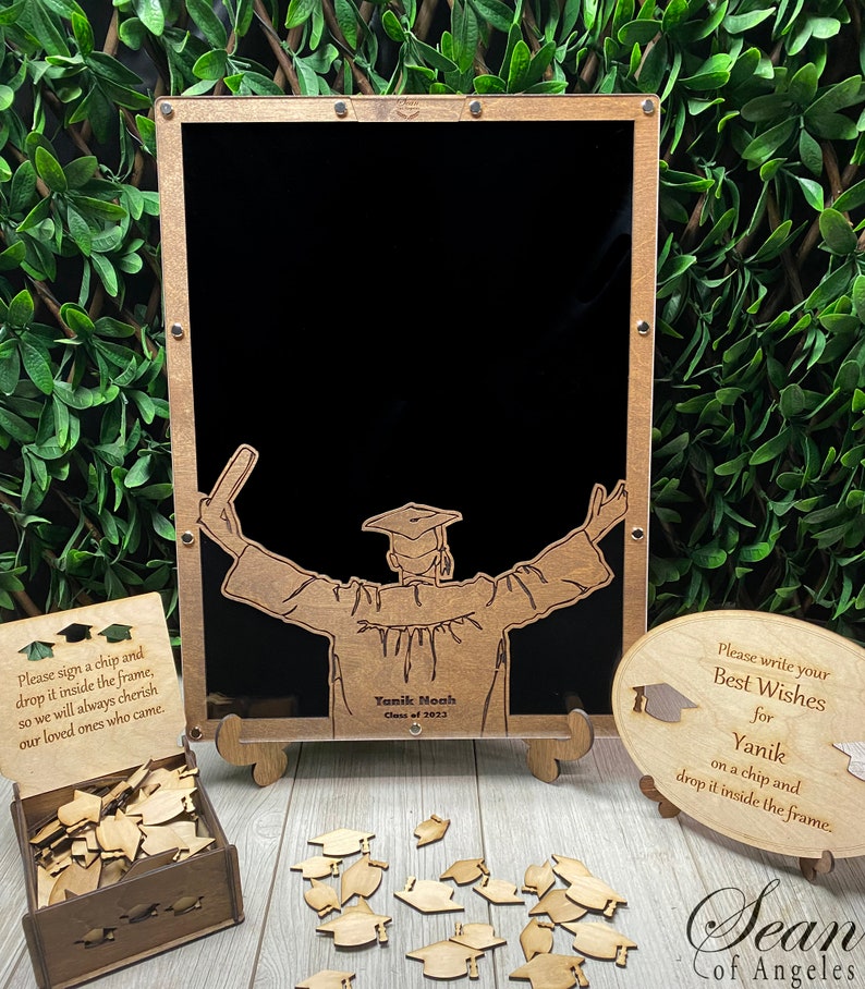Op de afbeelding: Houten bewaardoos voor afstuderen met een zwart schoolbordframe, een bord met de tekst "Schrijf je beste wensen voor Yanik op een chip en laat het in het frame vallen." en een klein houten doosje gevuld met houten chips in de vorm van afstudeerhoeden.