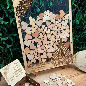 May include: Wooden Quincea&ntilde;era guest book alternative with a clear acrylic front, filled with wooden hearts. The frame features laser-cut roses and a dress design. Includes a wooden box and sign with the text "Please sign a heart and drop it inside the frame."