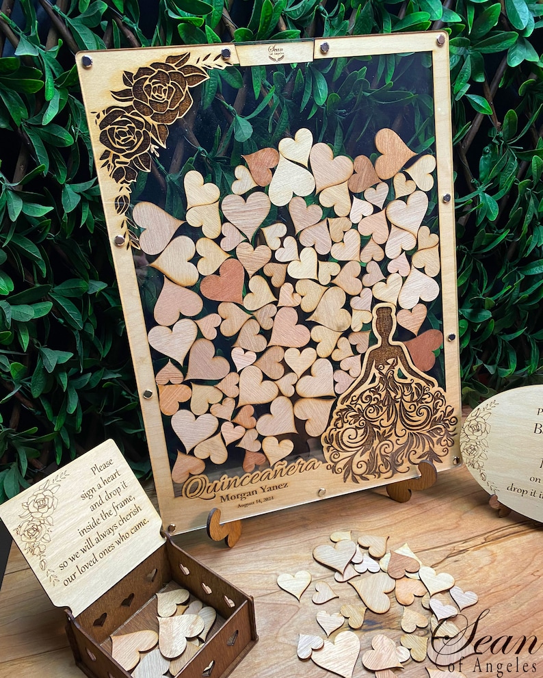 May include: Wooden Quincea&ntilde;era guest book alternative with a clear acrylic front, filled with wooden hearts. The frame features laser-cut roses and a dress design. Includes a wooden box and sign with the text "Please sign a heart and drop it inside the frame."