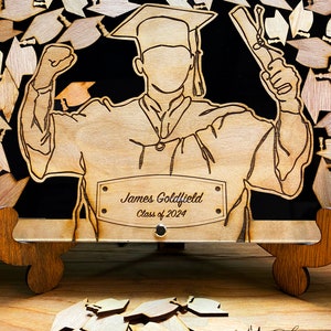 K&ouml;nnte beinhalten: Holzhalter f&uuml;r Abschlusskappen mit einer Aussparung eines Absolventen, der ein Diplom h&auml;lt. Der Halter hat eine personalisierte Namensschilder mit der Aufschrift "James Goldfield Klasse von 2024".